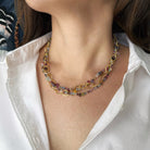 Layered necklaces with multicolored gems and gold chain worn by a person in a white shirt.