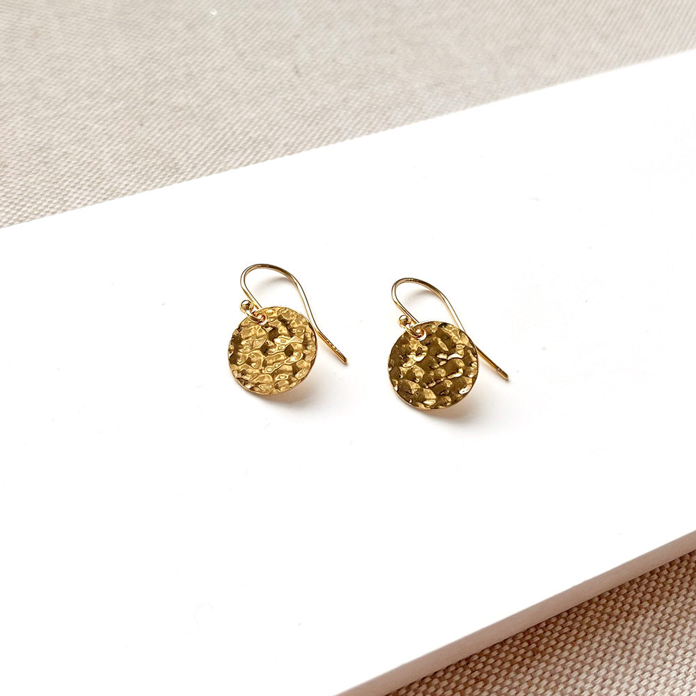A pair of gold-toned dangle earrings with textured circular discs, displayed on a white rectangular surface against a neutral beige fabric background.