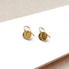 A pair of gold-toned dangle earrings with textured circular discs, displayed on a white rectangular surface against a neutral beige fabric background.