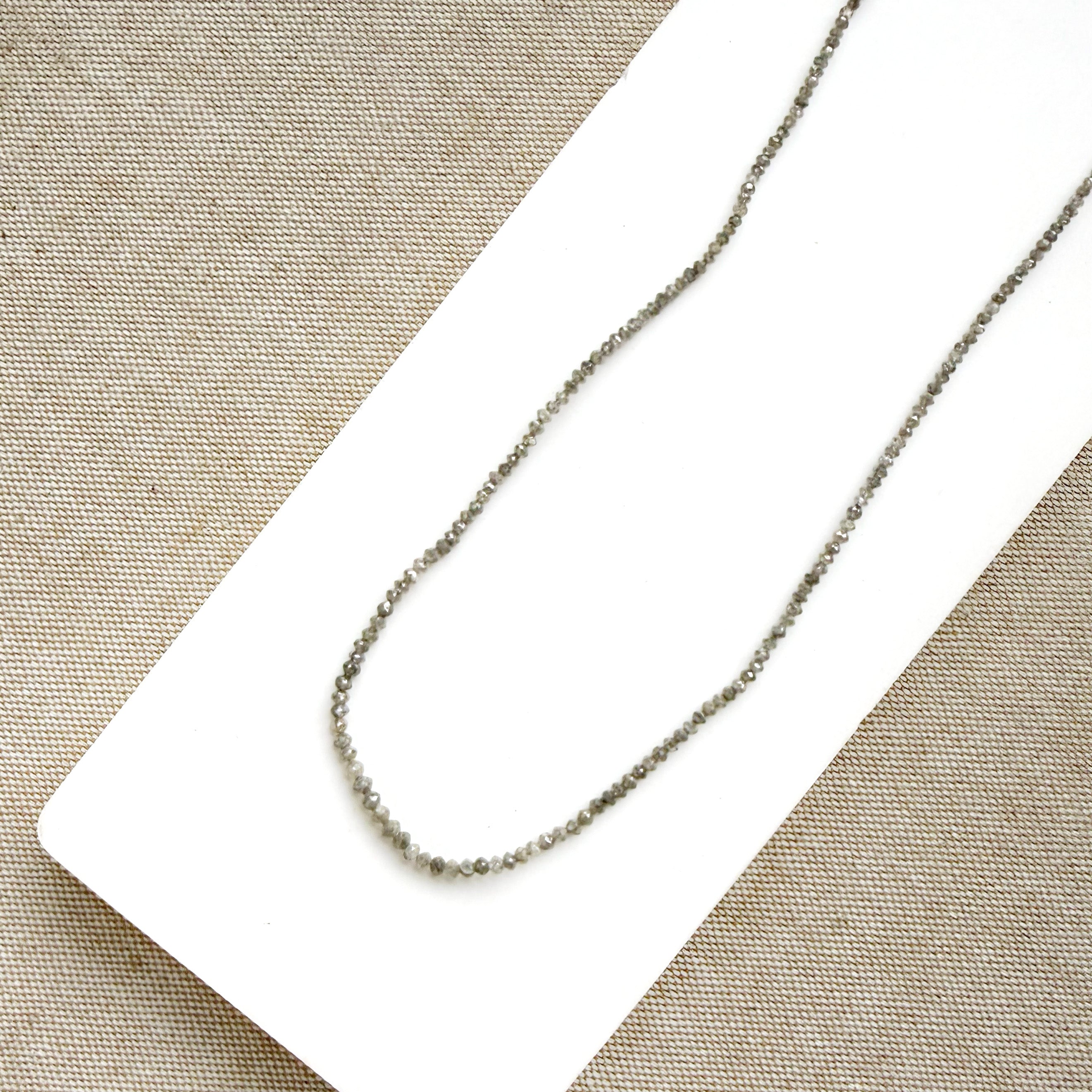A delicate rough cut diamond strung necklace draped over a white rectangular display card, set against a textured beige fabric background.