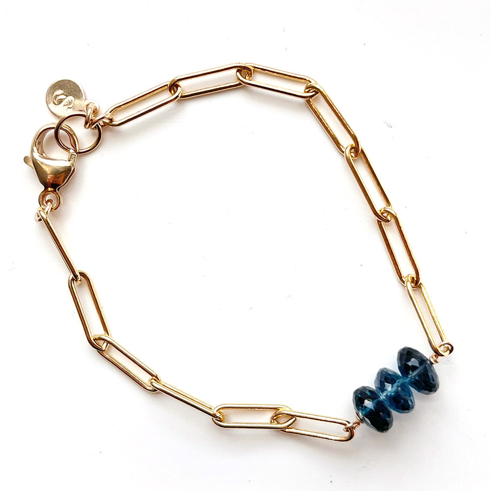 A gold paperclip chain bracelet with a small cluster of London Blue Topaz faceted beads, shown on a plain white background.