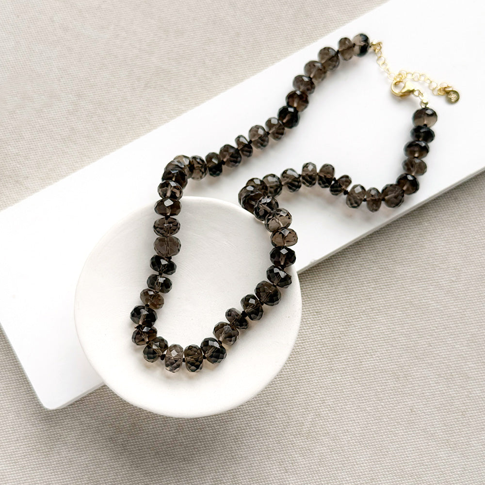 A knotted necklace of faceted smokey quartz gemstones resting in a white dish. The necklace features a gold-tone lobster clasp and extender chain.