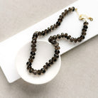A knotted necklace of faceted smokey quartz gemstones resting in a white dish. The necklace features a gold-tone lobster clasp and extender chain.