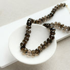 A knotted necklace of faceted smokey quartz gemstones resting on a white dish. The necklace features a gold-tone lobster clasp and extender chain.