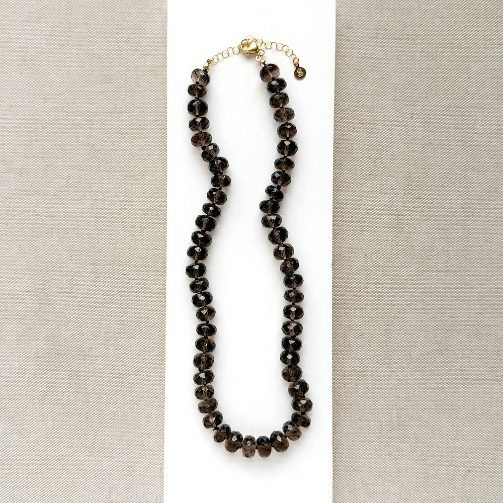 A knotted necklace of faceted smokey quartz gemstones resting on a white card. The necklace features a gold-tone lobster clasp and extender chain.