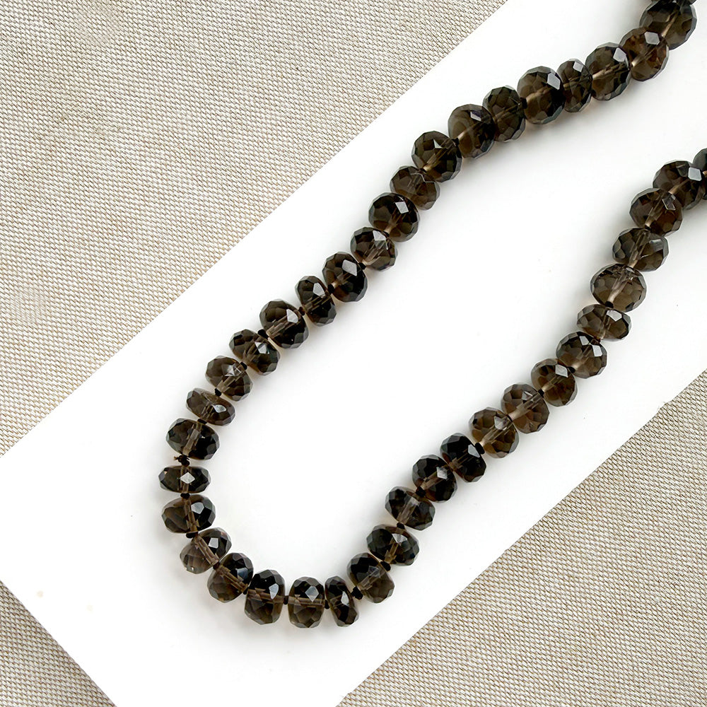 A knotted necklace of faceted smokey quartz gemstones resting on a white dish. The necklace features a gold-tone lobster clasp and extender chain.