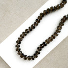 A knotted necklace of faceted smokey quartz gemstones resting on a white dish. The necklace features a gold-tone lobster clasp and extender chain.