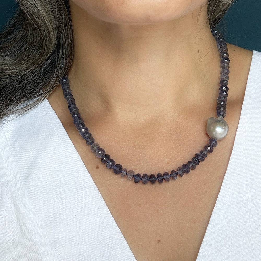 A close-up of a woman wearing a beaded necklace with faceted dark gray beads and a single large gray pearl, shown against her neck and white top.