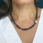 A close-up of a woman wearing a beaded necklace with faceted dark gray beads and a single large gray pearl, shown against her neck and white top.