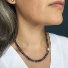 A close-up of a woman wearing a beaded necklace made of dark gray faceted beads with a single irregular gray pearl accent, paired with a white top.