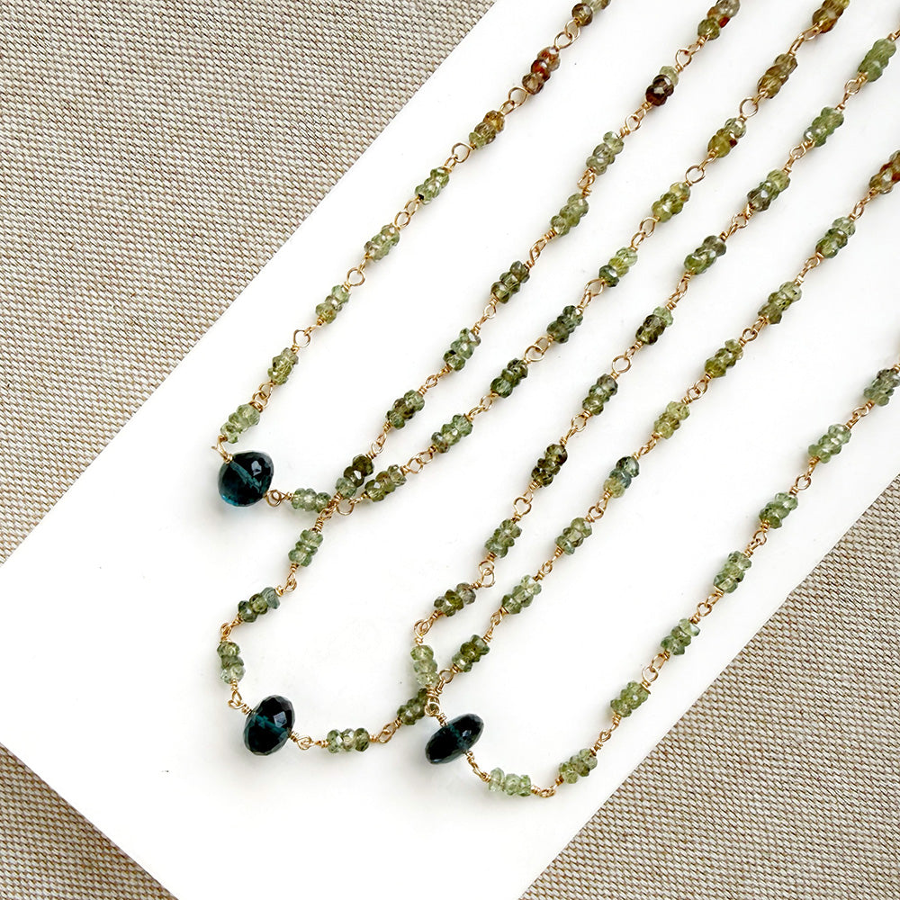 A necklace featuring small green faceted sapphires accented with a lager London Blue Topaz gemstone, displayed on a white rectangular card over a textured beige fabric background.