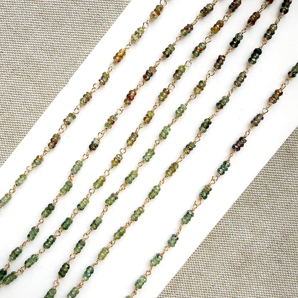 A necklace featuring small green faceted sapphires accented with a lager London Blue Topaz gemstone, displayed on a white rectangular card over a textured beige fabric background.