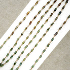 A necklace featuring small green faceted sapphires accented with a lager London Blue Topaz gemstone, displayed on a white rectangular card over a textured beige fabric background.