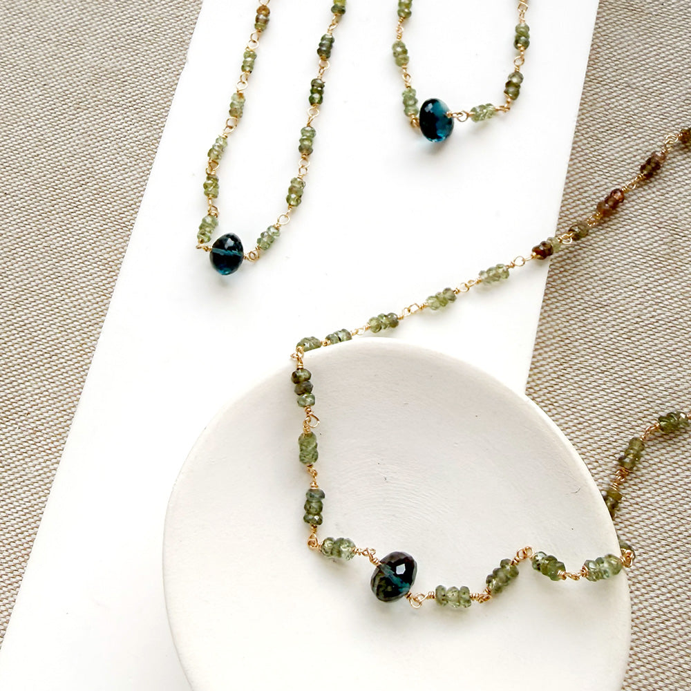 A necklace featuring small green faceted sapphires accented with a lager London Blue Topaz gemstone, displayed on a white rectangular card over a textured beige fabric background.