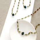 A necklace featuring small green faceted sapphires accented with a lager London Blue Topaz gemstone, displayed on a white rectangular card over a textured beige fabric background.
