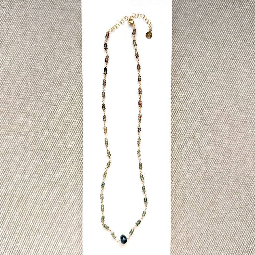 A necklace featuring small green faceted sapphires accented with a lager London Blue Topaz gemstone, displayed on a white rectangular card over a textured beige fabric background.