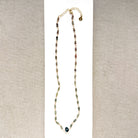 A necklace featuring small green faceted sapphires accented with a lager London Blue Topaz gemstone, displayed on a white rectangular card over a textured beige fabric background.