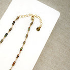 A necklace featuring small green faceted sapphires accented with a lager London Blue Topaz gemstone, displayed on a white rectangular card over a textured beige fabric background.