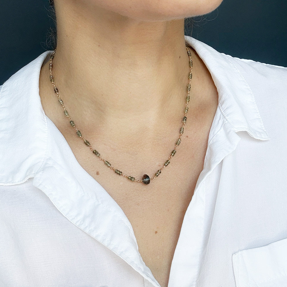 A necklace featuring small green faceted sapphires accented with a lager London Blue Topaz gemstone, displayed on a white rectangular card over a textured beige fabric background.