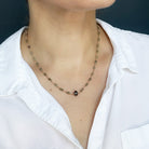 A necklace featuring small green faceted sapphires accented with a lager London Blue Topaz gemstone, displayed on a white rectangular card over a textured beige fabric background.