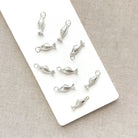 Nine small silver fish-shaped jewelry charms with textured scales and hanging loops, arranged on a white rectangular card over a beige fabric background.