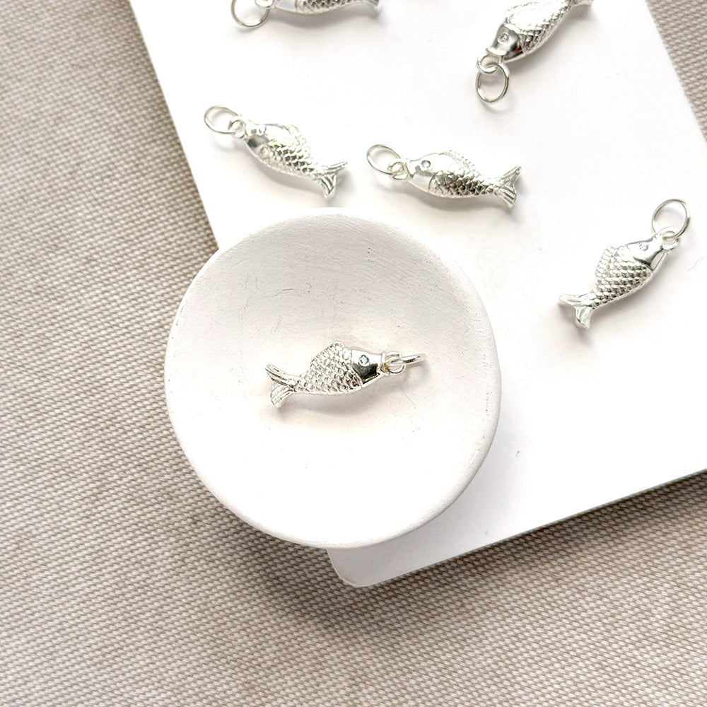 Silver fish-shaped jewelry charms displayed on a white card over a beige textured fabric background.