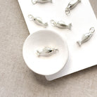 Silver fish-shaped jewelry charms displayed on a white card over a beige textured fabric background.