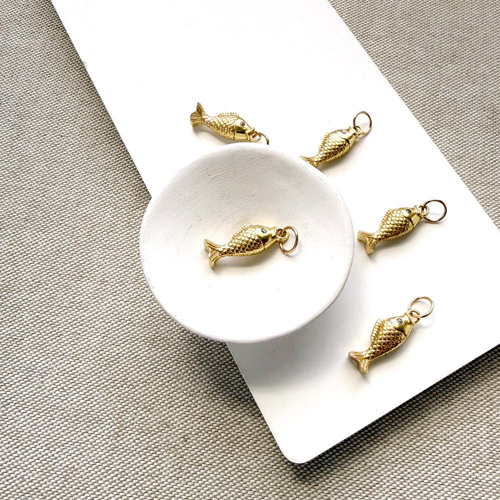 Five small gold fish-shaped charms with metal loops, arranged on a white rectangular board over a textured beige fabric background.
