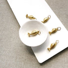 Five small gold fish-shaped charms with metal loops, arranged on a white rectangular board over a textured beige fabric background.