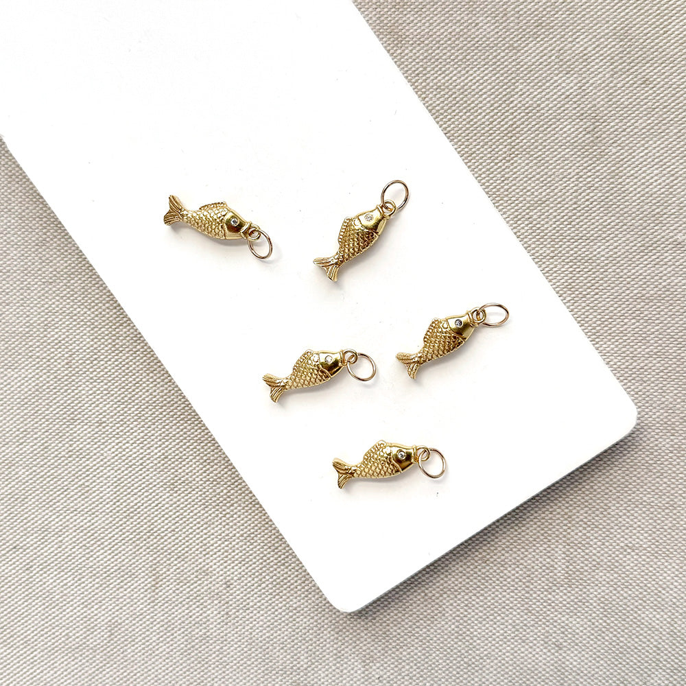 Five small gold fish-shaped jewelry charms with attached loops, arranged on a white rectangular card over a light beige textured fabric background.