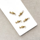 Five small gold fish-shaped jewelry charms with attached loops, arranged on a white rectangular card over a light beige textured fabric background.