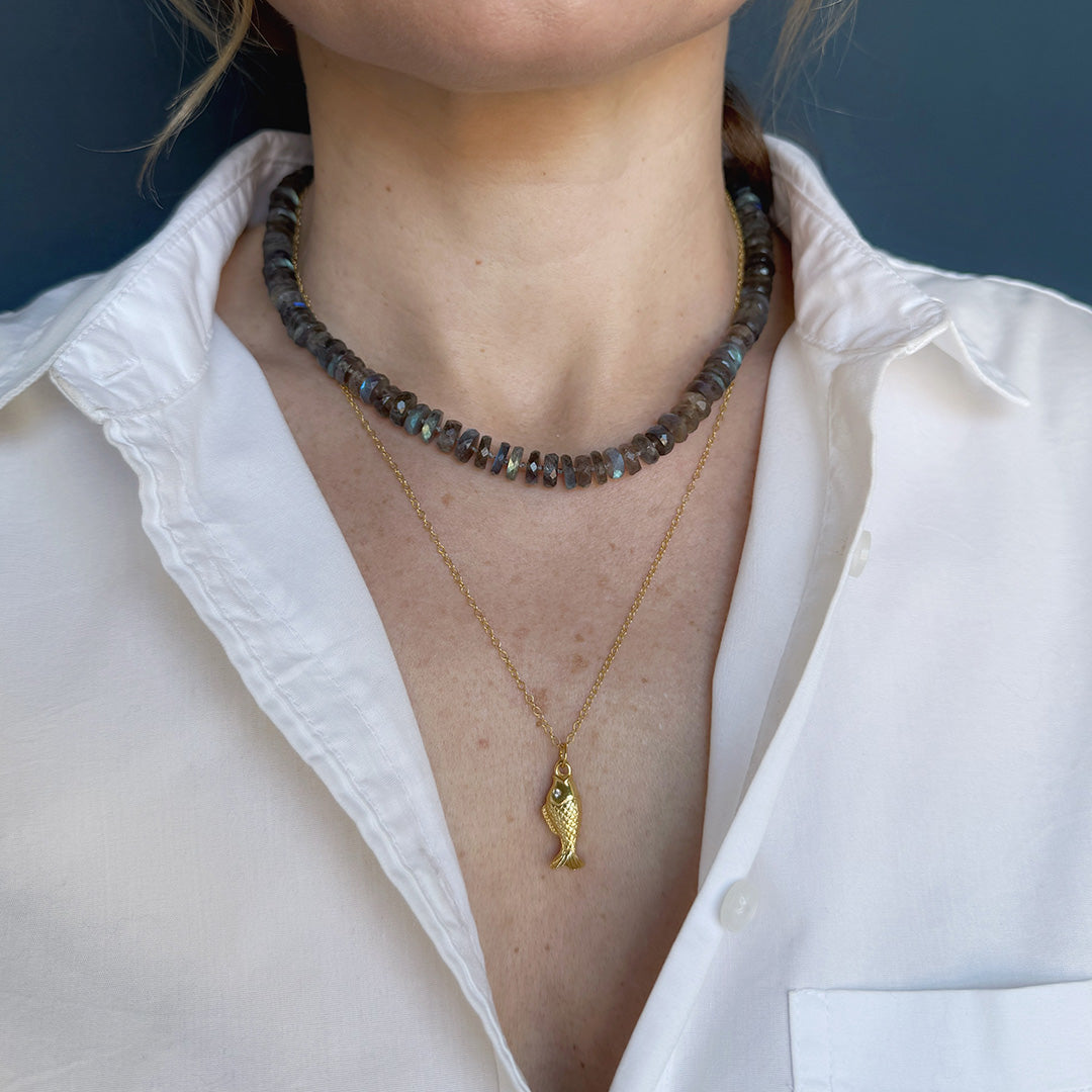 Person wearing two necklaces, one with a fish pendant, against a white shirt.