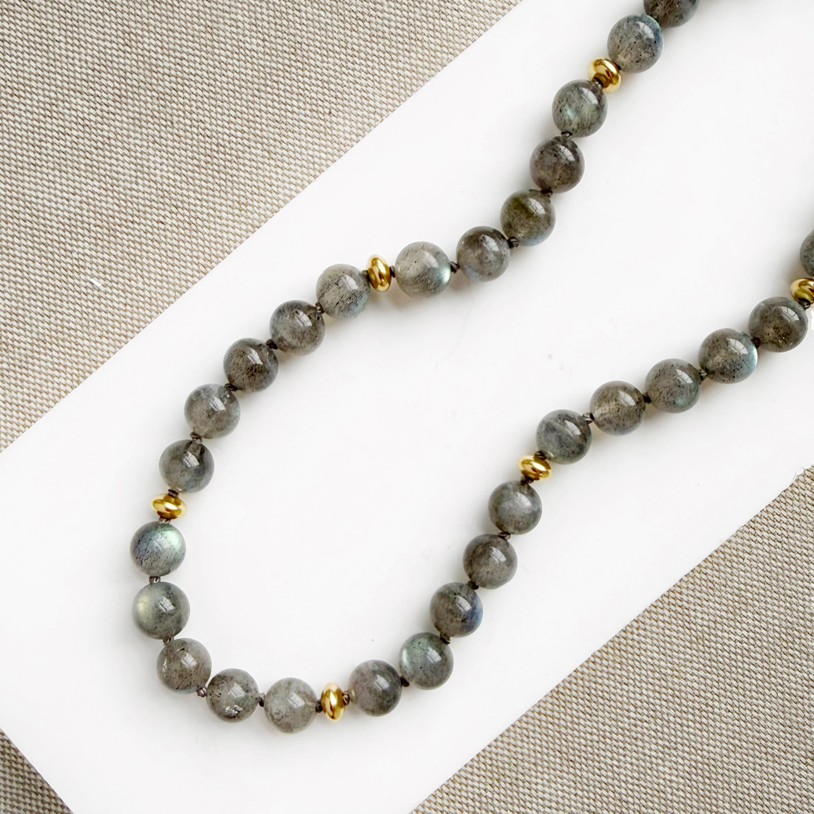 A necklace with round labradorite gemstones, featuring gold disc bead accents, knotted on silk, displayed on a white surface.