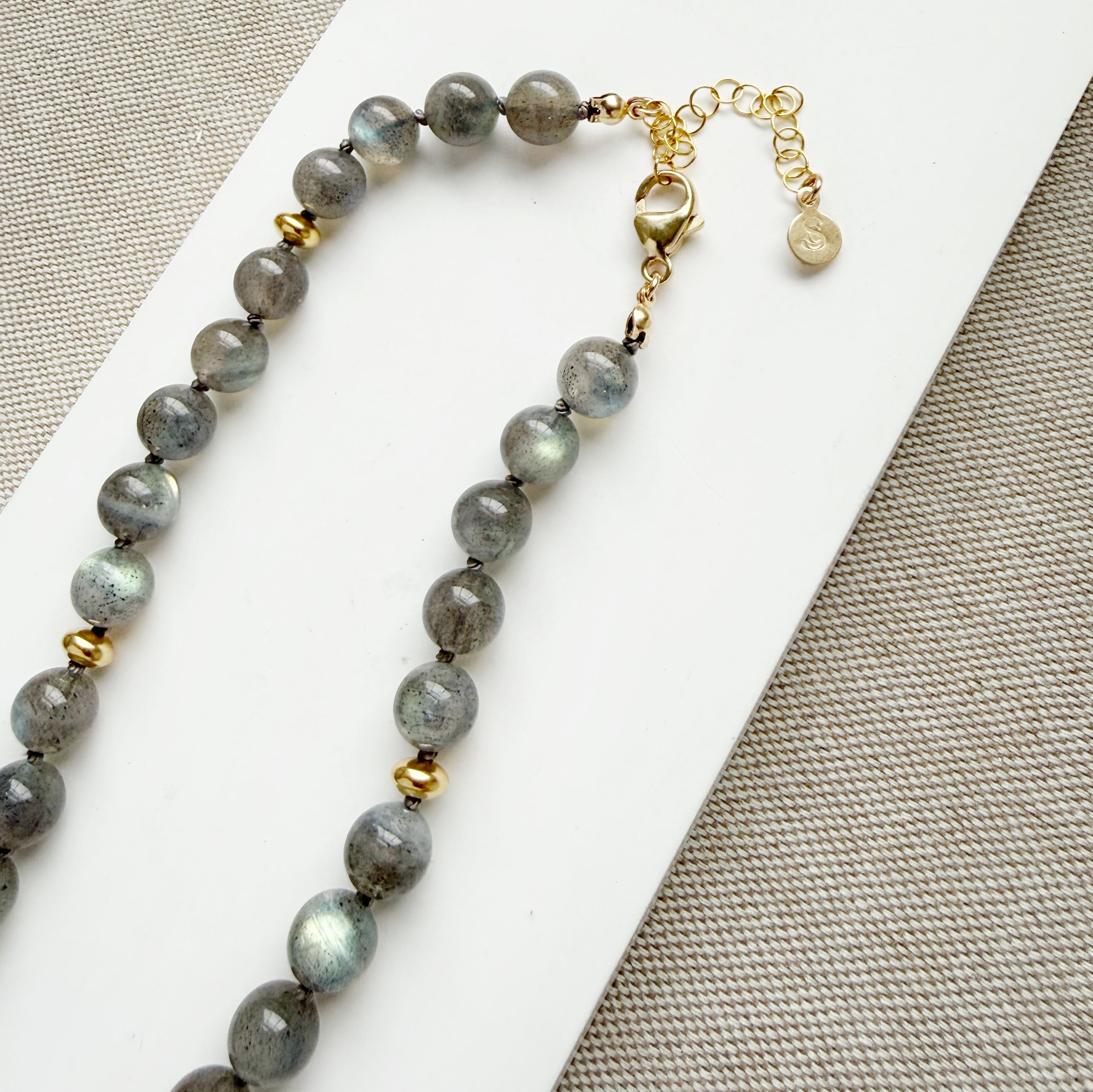 Clasp detail of a necklace with round labradorite gemstones, featuring gold disc bead accents, knotted on silk, displayed on a white surface.