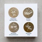 Four gold floral charms on a white card with month and flower name labels, on a textured beige background.