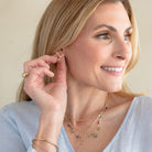 A close-up lifestyle shot of a woman wearing delicate gold jewelry, including a small gold hoop earring, layered gold necklaces, and a gold ring, against a soft beige background.