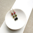 A pair of gold earrings with watermelon tourmaline beads, displayed on a small white ceramic dish over a beige woven fabric background.