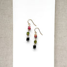 A pair of gold earrings with watermelon tourmaline gemstones, displayed on a white card between textured beige fabric borders.