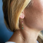 A close-up lifestyle shot of a woman's ear wearing a delicate gold dangle earring watermelon tourmaline gemstone beads, alongside a thin gold chain necklace.