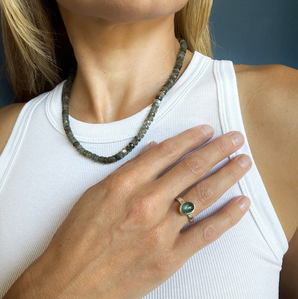 A close-up of a woman wearing a chunky natural stone necklace and a gold ring with a green oval stone, wearing a white ribbed tank top.