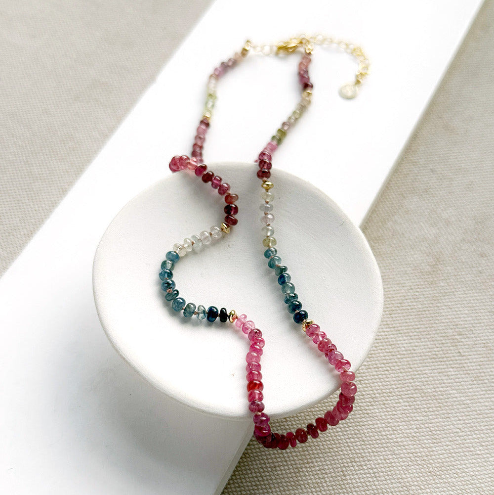 A sapphire necklace draped over a small white dish, featuring multicolored faceted beads transitioning through pink, red, blue, green, and clear tones, finished with a gold clasp.