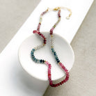 A sapphire necklace draped over a small white dish, featuring multicolored faceted beads transitioning through pink, red, blue, green, and clear tones, finished with a gold clasp.