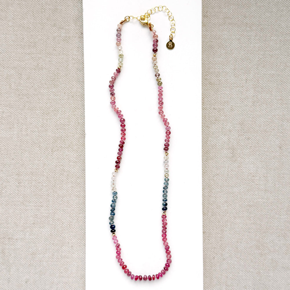 A sapphire necklace on a white card, featuring multicolored faceted beads transitioning through pink, red, blue, green, and clear tones, finished with a gold clasp.