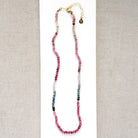 A sapphire necklace on a white card, featuring multicolored faceted beads transitioning through pink, red, blue, green, and clear tones, finished with a gold clasp.