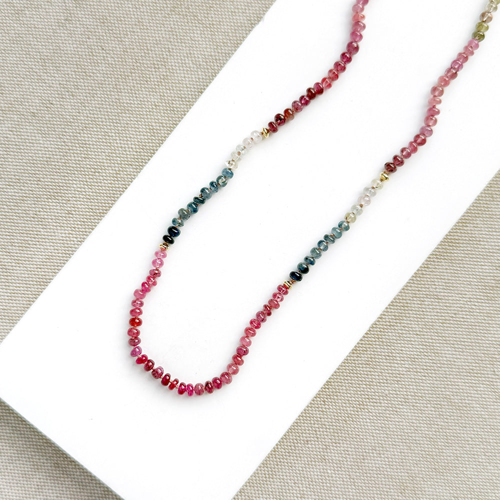 A sapphire necklace on a white card, featuring multicolored faceted beads transitioning through pink, red, blue, green, and clear tones, finished with a gold clasp.