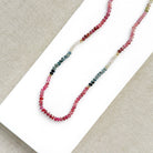 A sapphire necklace on a white card, featuring multicolored faceted beads transitioning through pink, red, blue, green, and clear tones, finished with a gold clasp.