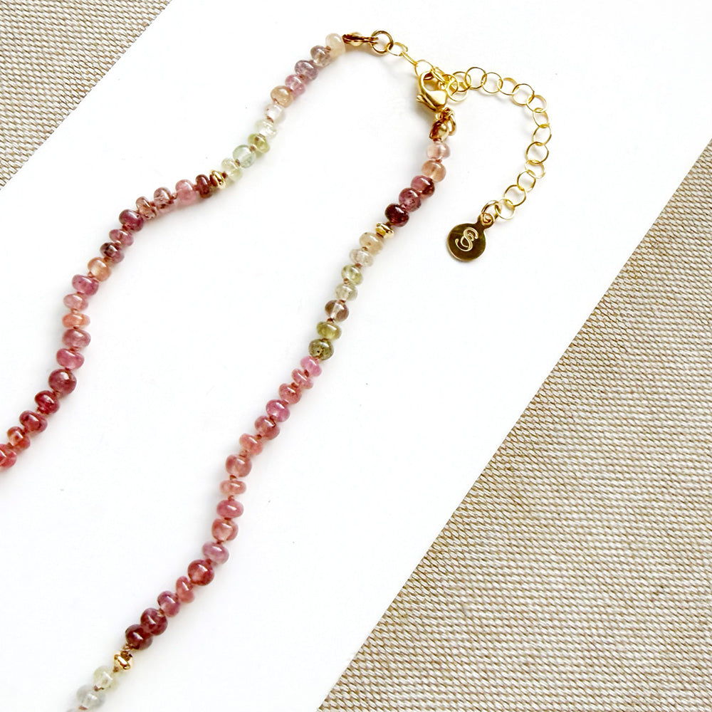 A sapphire necklace on a white card, featuring multicolored faceted beads transitioning through pink, red, blue, green, and clear tones, finished with a gold clasp.