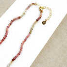 A sapphire necklace on a white card, featuring multicolored faceted beads transitioning through pink, red, blue, green, and clear tones, finished with a gold clasp.