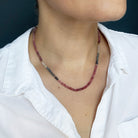 A close-up of a person wearing a beaded necklace with pinkish-red, gray, and clear sapphires, styled with a white collared shirt against a dark background.