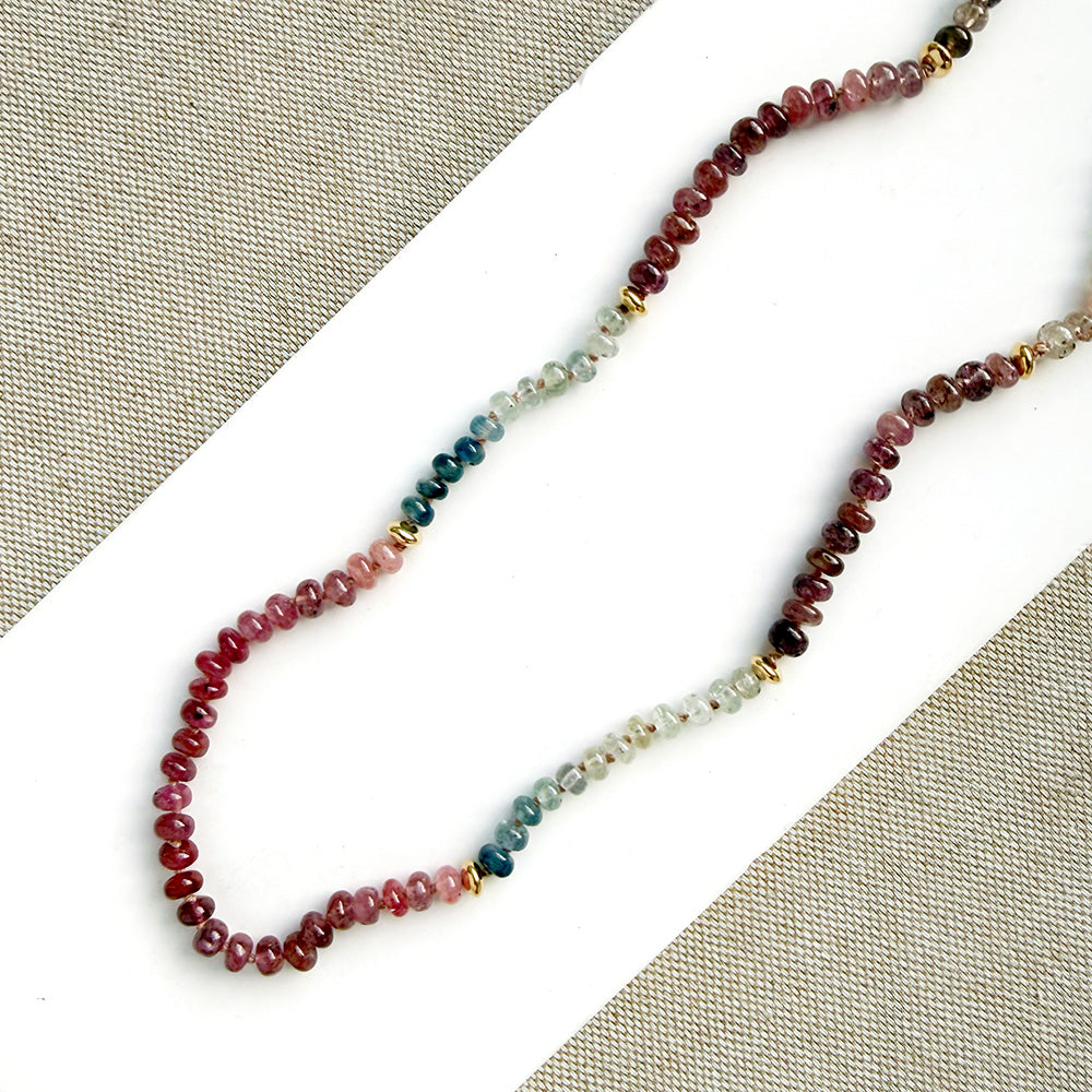 A beaded strand necklace laid diagonally on a white surface with textured beige fabric in the corners. The necklace features small faceted beads transitioning through red, pink, teal, green, and clear/light gray, separated by small gold spacer beads.